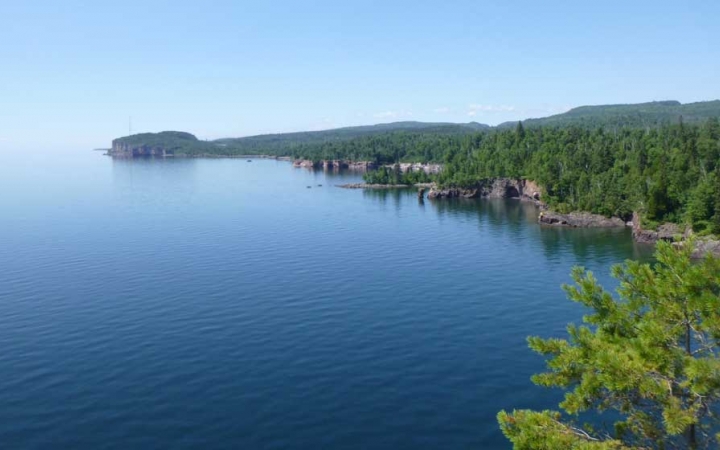 The rocky and tree-lined shore of Lake Superior frames the calm, blue water of the lake. 